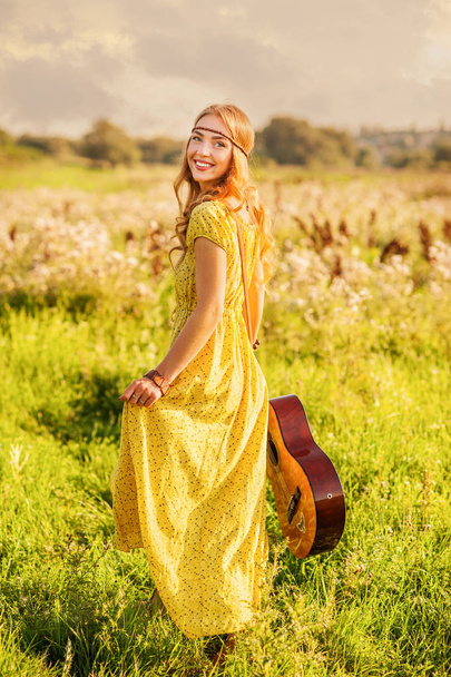 Menina feliz em estilo boêmio vestido amarelo com guitarra no campo virou-se para trás ao pôr do sol
 - Foto, Imagem