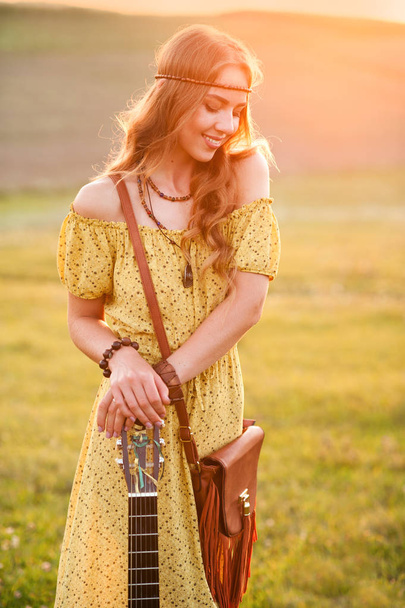 κορίτσι σε κίτρινο φόρεμα μποέμικο στυλ, κρατώντας μια κιθάρα στο γήπεδο με ένα ζεστό και άνετο ηλιοβασίλεμα ατμόσφαιρα - Φωτογραφία, εικόνα