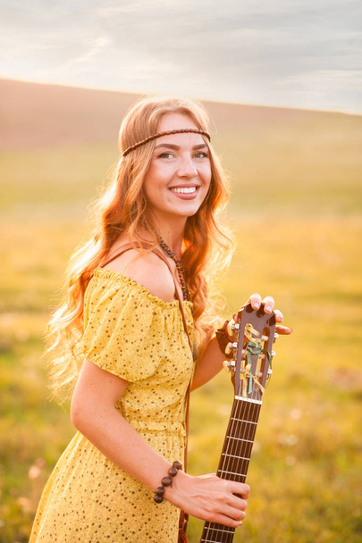 girl in yellow dress bohemian style, holding a guitar on the field with a warm and comfortable atmosphere sunset - Photo, Image