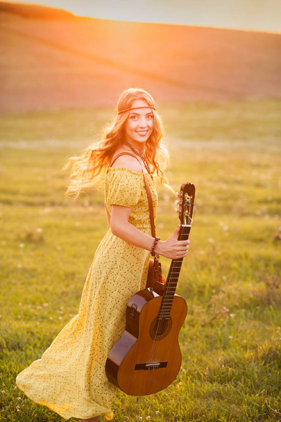 Beautiful hippie girl with guitar dancing on field at sunset - 写真・画像