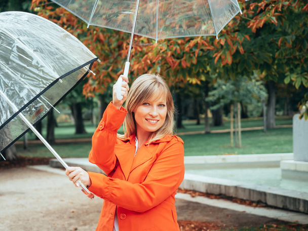 Biała dziewczyna w czerwonym gabardynie bawi się dwoma przezroczystymi parasolami w parku - Zdjęcie, obraz