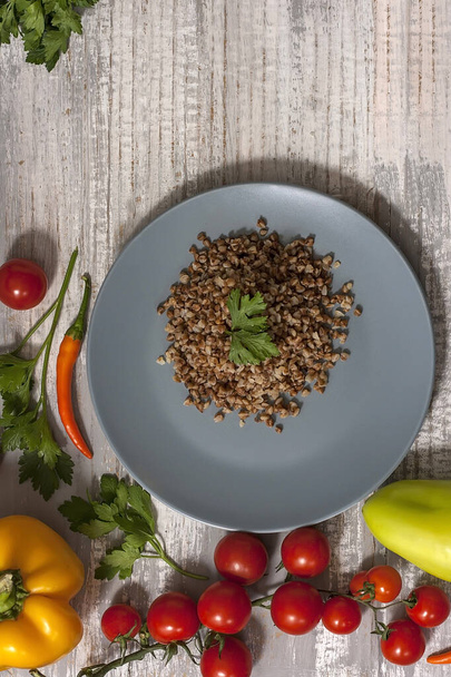 Cooked buckwheat porridge in a grey plate on a light white wooden background with vegetables. - Photo, Image