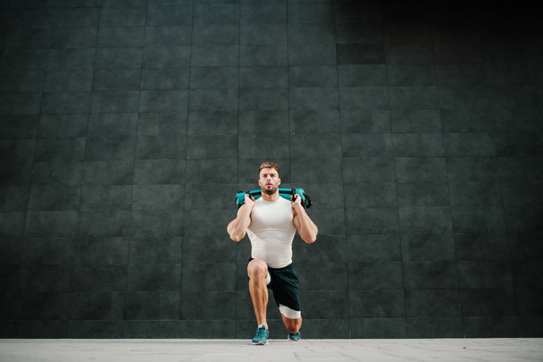 Front view of handsome muscular bearded caucasian man in shorts and t-shirt holding training bag while doing lunges. - Photo, Image