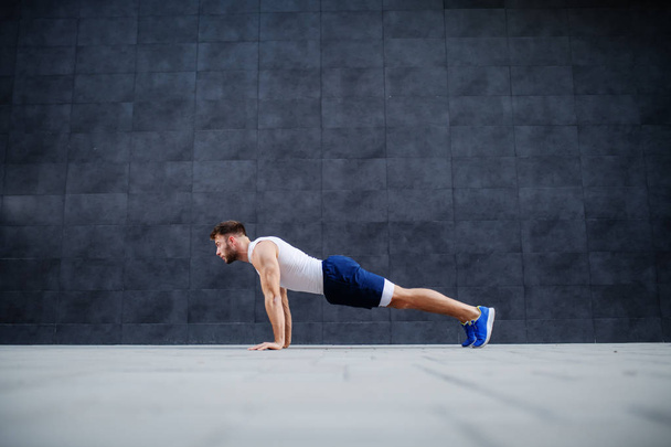 Side view of handsome Caucasian muscular bearded man in shorts and t-shirt doing push-ups. In background is gray wall. - Fotoğraf, Görsel