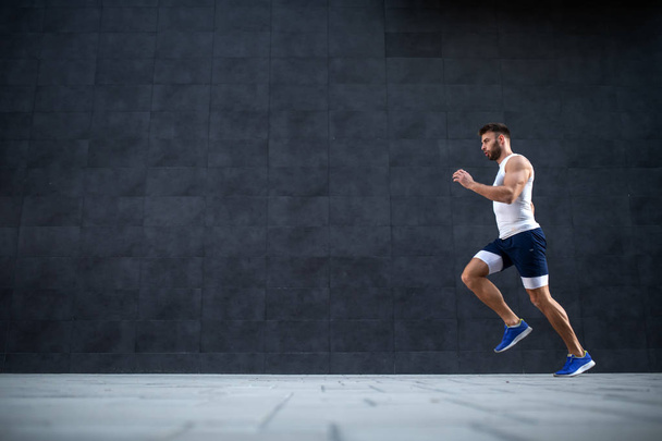 Pohled z boku na pohledného bělocha, svalnatého muže běžícího rychle ven. V pozadí je šedá zeď. - Fotografie, Obrázek
