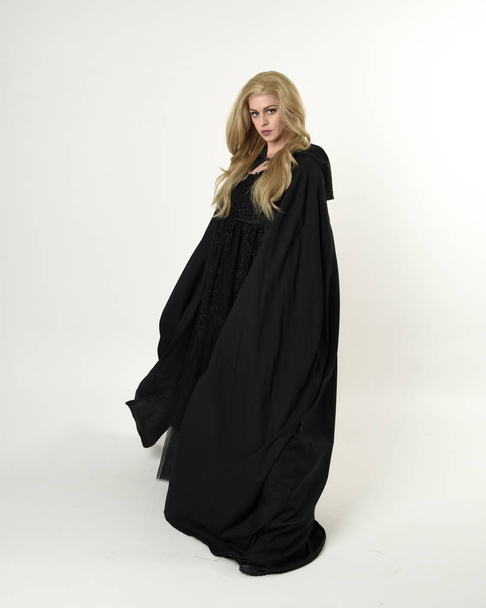 Ganzkörperporträt eines blonden Mädchens, das einen langen schwarzen, fließenden Mantel trägt, in stehender Pose mit weißem Studiohintergrund. - Foto, Bild