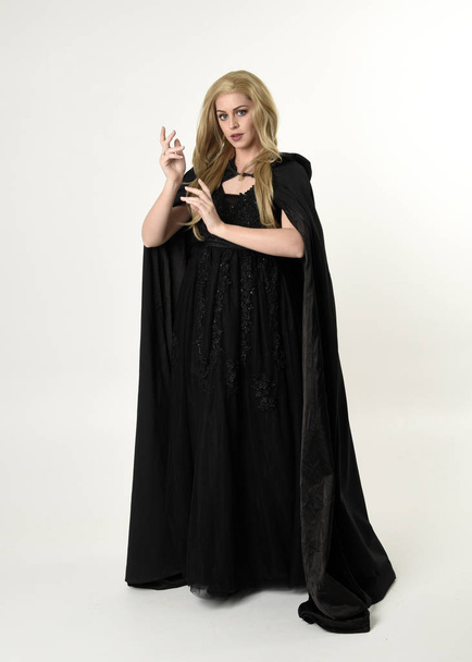 full length portrait of blonde girl wearing long black flowing cloak, standing pose  with  a white studio background. - Photo, Image