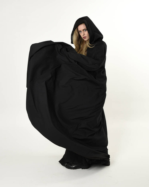 Ganzkörperporträt eines blonden Mädchens, das einen langen schwarzen, fließenden Mantel trägt, in stehender Pose mit weißem Studiohintergrund. - Foto, Bild