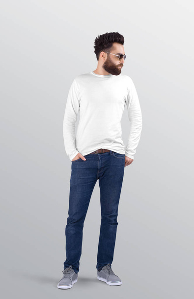 Uomo barbuto in piedi con girocollo bianco liscio camicia a maniche lunghe con pantaloni jeans blu denim. Fondo isolato
. - Foto, immagini