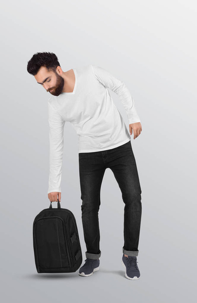 Standing male model wearing plain white v neck long sleeve shirt in black denim jeans pant, picking blag backpack. Isolated background - Фото, изображение