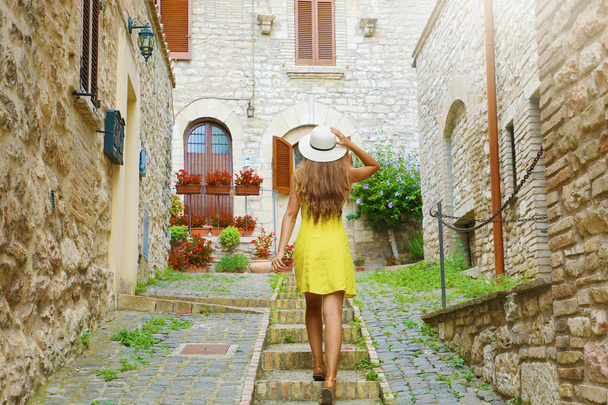 Mujer joven y curiosa con vestido amarillo y sombrero va arriba en la calle medieval en Toscana, Italia. Vista trasera de la alegre chica feliz visitando el centro de Italia
. - Foto, imagen