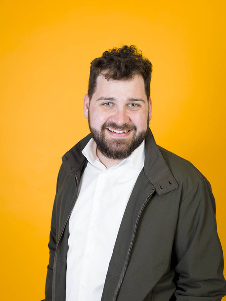 emotional man with black beard and black hair in white shirt and black jacket smiling and looking at camera on studio orange background  - 写真・画像