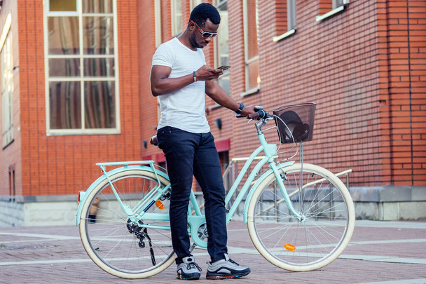 handsome African American man stylish well-dressed standing with a blue bicycle and holding phone in hand red brick building background . sports ecological transport concept - Fotografie, Obrázek