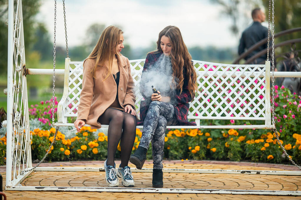 Smoking and vaping. Young beautiful white teeage girls in casual clothes sit on a vintage bench and smoke and vape cigarettes on the street in the evening. Electronic cigarette. Bad habit. - Foto, immagini