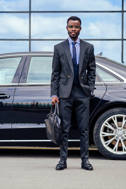 successful businessman handsome African American man in a stylish suit in gray jacket and glasses standing in front of a cool new black car on the street - Фото, зображення
