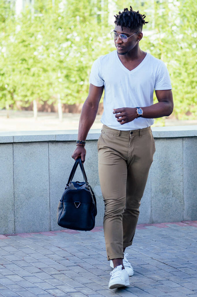 stylish and handsome African American man with cool dreadlocks in brown pants and a white T-shirt fashion look holding the bag walking on the street - Foto, Bild