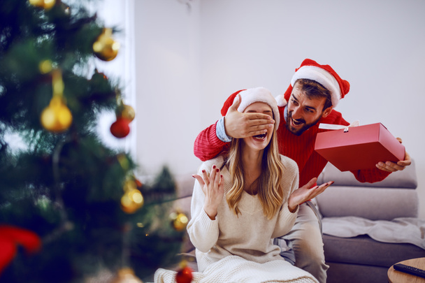 Smiling handsome bearded caucasian man covering his girlfriend's eyes and holding gift. Woman sitting on sofa. Both having santa hats on heads. Living room interior. - Photo, image