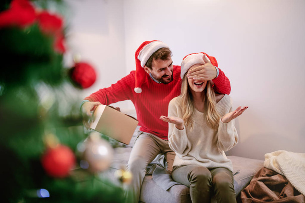 Smiling handsome bearded caucasian man covering his girlfriend's eyes and hiding gift behind back. Woman sitting on sofa. Both having santa hats on heads. Living room interior. - Photo, Image