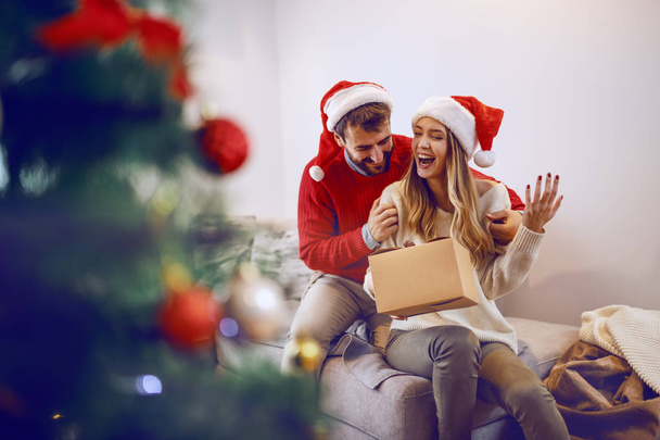 Excited caucasian smiling woman holding gift and sitting on sofa in living room. Her boyfriend hugging her. Both having santa hats on heads. In foreground is fir. Living room interior. - Photo, Image