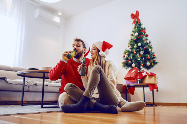 Αξιολάτρευτο ζευγάρι Καυκάσιων με καπέλα Σάντα σε κεφάλια που κάθονται στο πάτωμα του σαλονιού και πίνουν μπύρα. Στο βάθος είναι χριστουγεννιάτικο δέντρο με δώρα. - Φωτογραφία, εικόνα