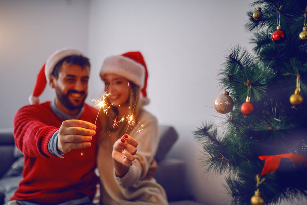 Happy handsome caucasian couple with santa hats on heads sitting on sofa in living room,celebrating New Year and holding sparklers. In foreground is christmas tree. Selective focus on sparklers. - Foto, Imagem