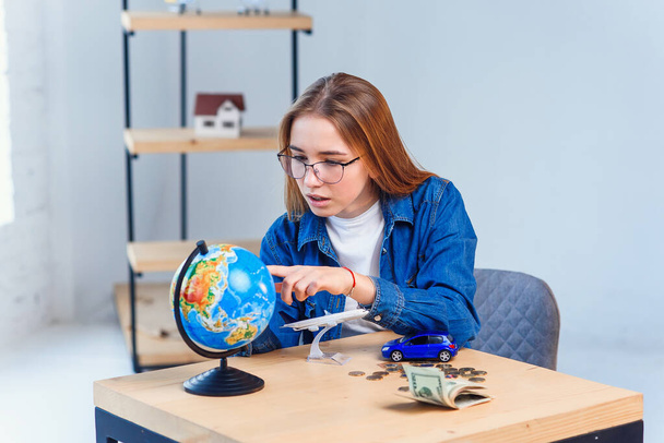 Young caucasian woman sits at the table and rotates the globe to choose the country for vacation and make notes in the note book. - Foto, imagen