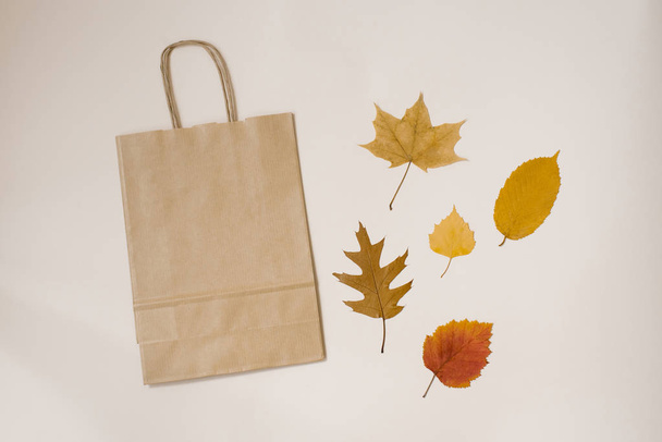 Бумажный мешок с ручками для покупок и осенью опавшие листья желтого и красного цвета на бежевом фоне. Осенние скидки, падение цен, черная пятница, продажа
 - Фото, изображение