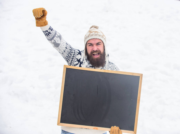 Winter event. Man with beard hold chalkboard copy space. Check it out. Winter holidays. Emotional guy snowy nature background. Winter announcement. Hipster knitted hat and gloves show blackboard - Фото, зображення