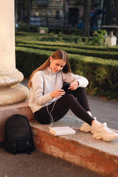 Joven hermosa chica estudiante con estilo en suéter acogedor de ensueño utilizando el teléfono celular durante el descanso de estudio al aire libre
  - Foto, imagen