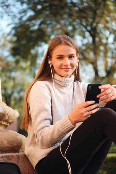 Молода красива студентка в затишному светрі з мобільним телефоном, щасливо дивлячись в камеру під час перерви навчання на відкритому повітрі
  - Фото, зображення