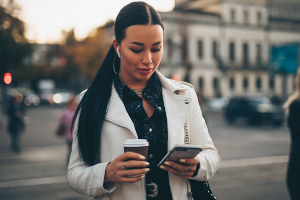 Portrait of woman using mobile phone while holding coffee cup on a city street - Photo, Image