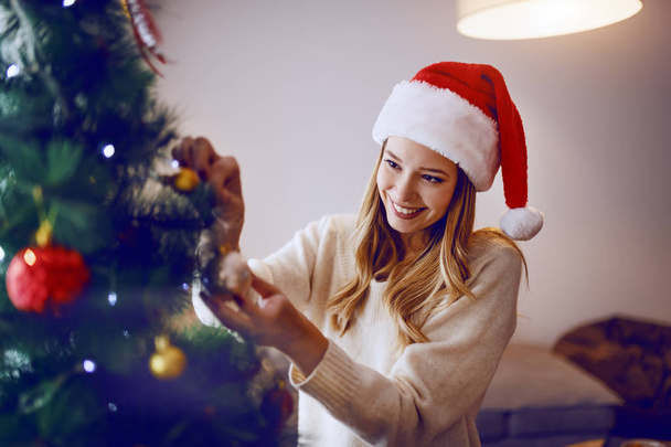 Superbe femme blonde caucasienne souriante avec chapeau de Père Noël sur la tête debout dans le salon et décorant l'arbre de Noël
. - Photo, image