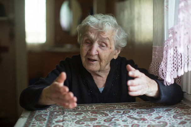 Emotional elderly woman tells gesturing sitting at a table in the house. - Photo, Image