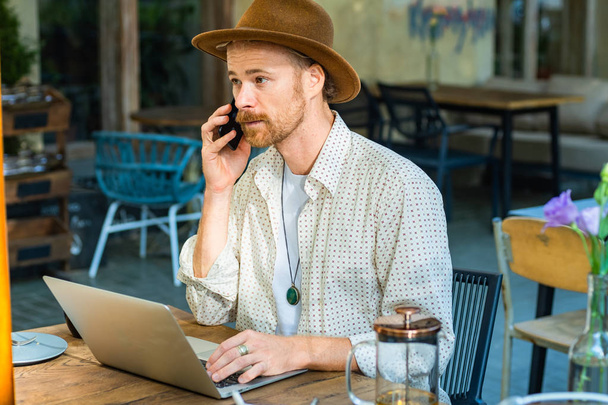 Young stylish hipster in hat work with laptop outdoors in the street cafe   - Foto, Imagen