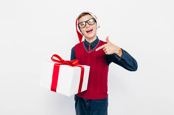 Happy boy in a red Santa hat. Stylish boy with a Christmas gift, joyfully pointing his finger at the gift, on a white background - Photo, image