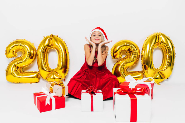 Happy little girl in Santa hat, stylish baby sitting with Christmas gifts sitting on white background with gold 2020 numbers - Fotó, kép
