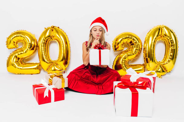 Pensive little girl in Santa hat, stylish baby sitting pensive with Christmas gifts sitting on white background with gold 2020 numbers - Foto, immagini