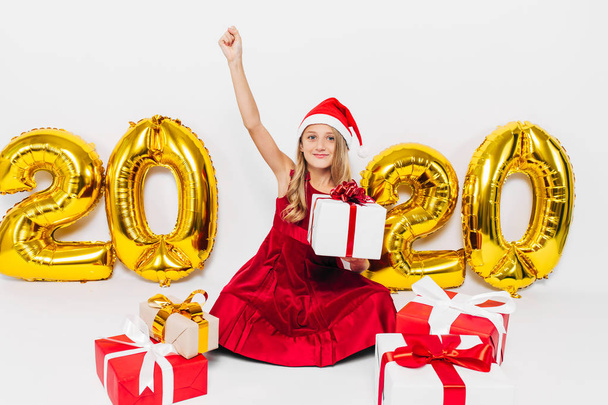 Happy little girl in Santa hat, stylish baby rejoices Christmas gift by showing victory gesture while sitting on white background with gold 2020 numbers - Foto, Imagen