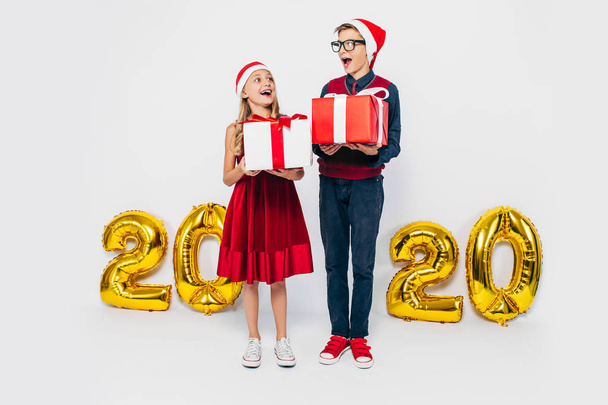 Happy little girl and boy wearing Santa hat, stylish brother and sister are rejoicing holding Christmas gifts, on white background with gold 2020 figures - Fotoğraf, Görsel