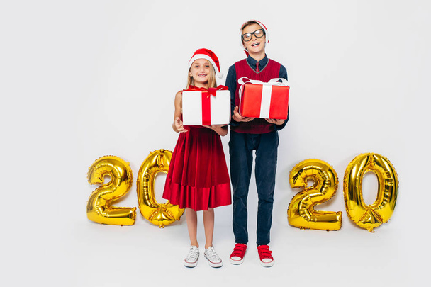 Happy little girl and boy wearing Santa hat, stylish brother and sister are rejoicing holding Christmas gifts, on white background with gold 2020 figures - Foto, Bild