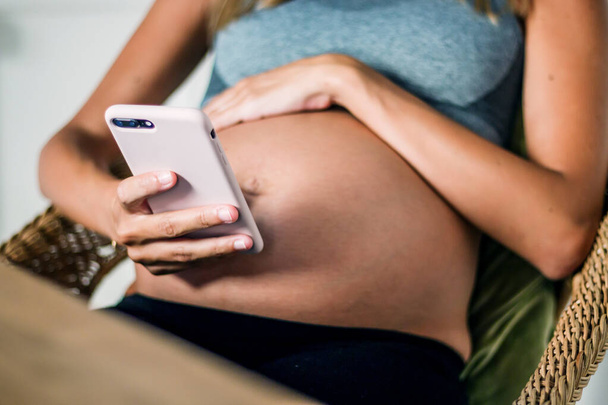 Lövés gyönyörű terhes fiatal nő használja a mobiltelefonját, miközben otthon pihen - Fotó, kép
