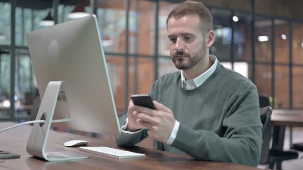 Fiatalember használja a mobilt, miközben az irodai asztalon ül - Felvétel, videó