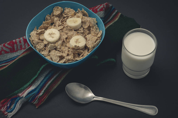 Cereal con leche y platanos sobre un mantel, con una cuchara y un vaso en fondo negro - Foto, Imagem