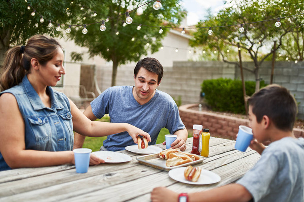 onnellinen latino perhe syö grillattuja hot dogeja piknikpöydällä takapihalla
 - Valokuva, kuva