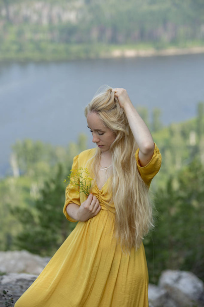 Beautiful young blonde girl with long hair in a yellow dress, look down, at the flower. Girl in nature - Fotografie, Obrázek