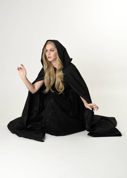 pełnowymiarowy portret blondynki noszącej długi czarny płaszcz, siedzącej na podłodze z białym tłem studio. - Zdjęcie, obraz