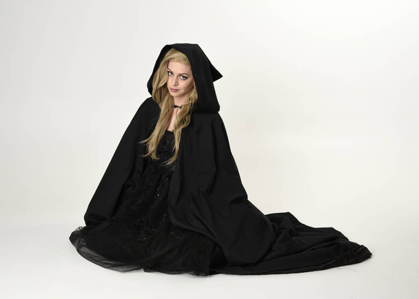 retrato de comprimento total de menina loira vestindo manto longo fluxo preto, sentado no chão com um fundo de estúdio branco
. - Foto, Imagem