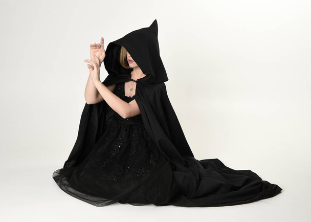 retrato de longitud completa de la chica rubia que lleva una larga capa de flujo negro, sentado en el suelo con un fondo de estudio blanco
. - Foto, Imagen
