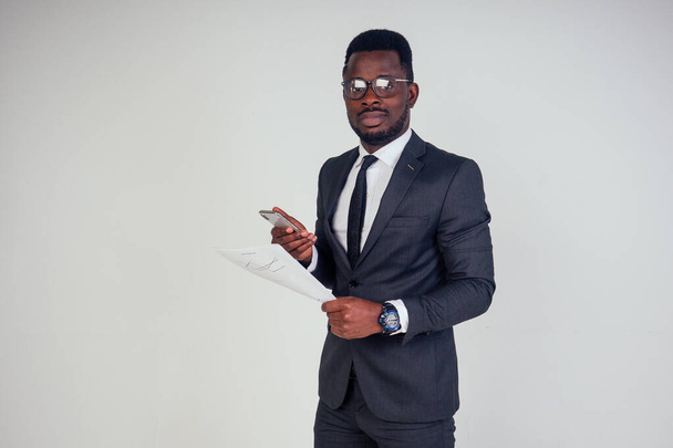 Handsome young African man in formal black suit holds a plastic paper cup with hot coffee cocoa tea to go and smartphone. businessman on coffee break after meeting on white background in studio - Foto, immagini