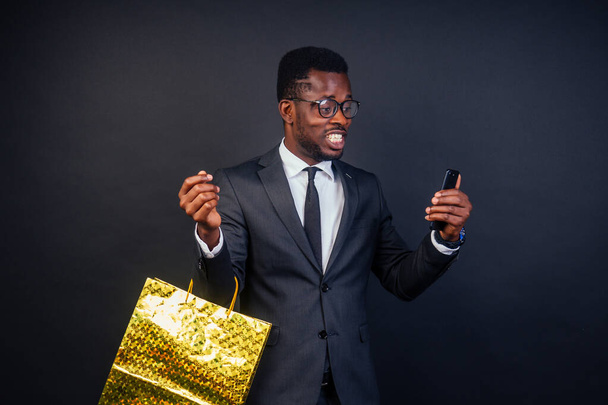 Kara Cuma indirim fikri. Cep telefonu kullanan Afro-Amerikan bir adam, mutlu esmer tenli iş adamı bir alışveriş çantası tutuyor. - Fotoğraf, Görsel